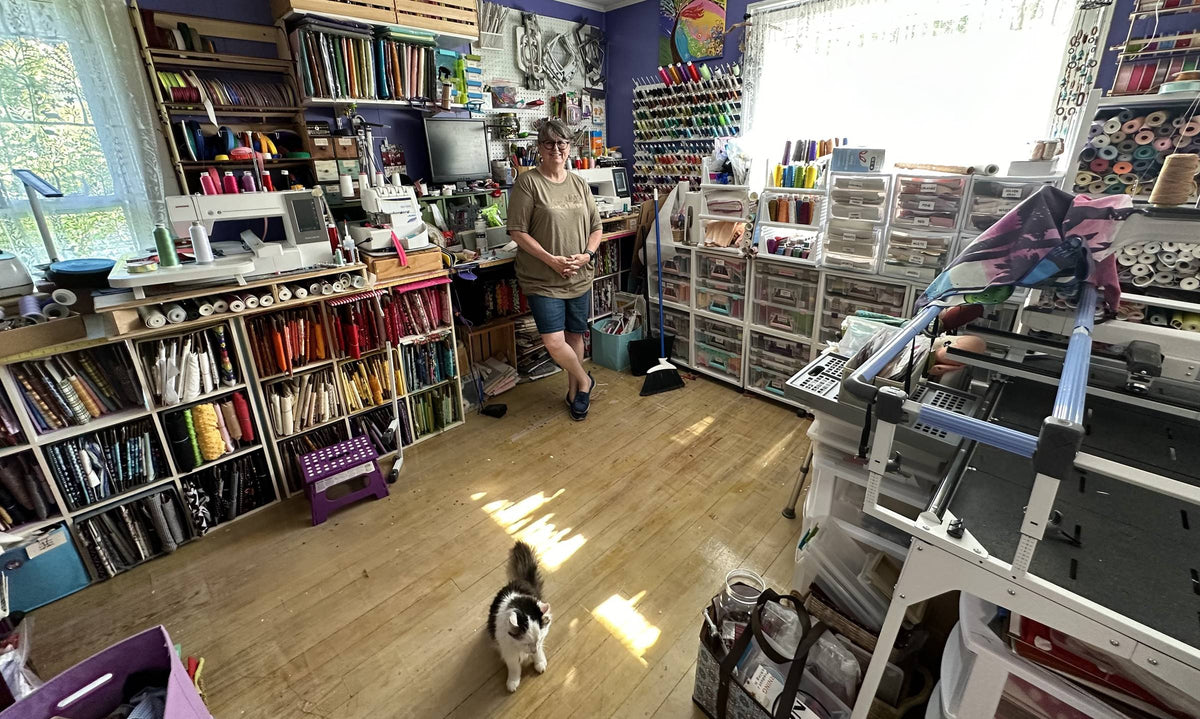 Kathy in her sewing studio with shelves of fabric, a wall of thread and her cat